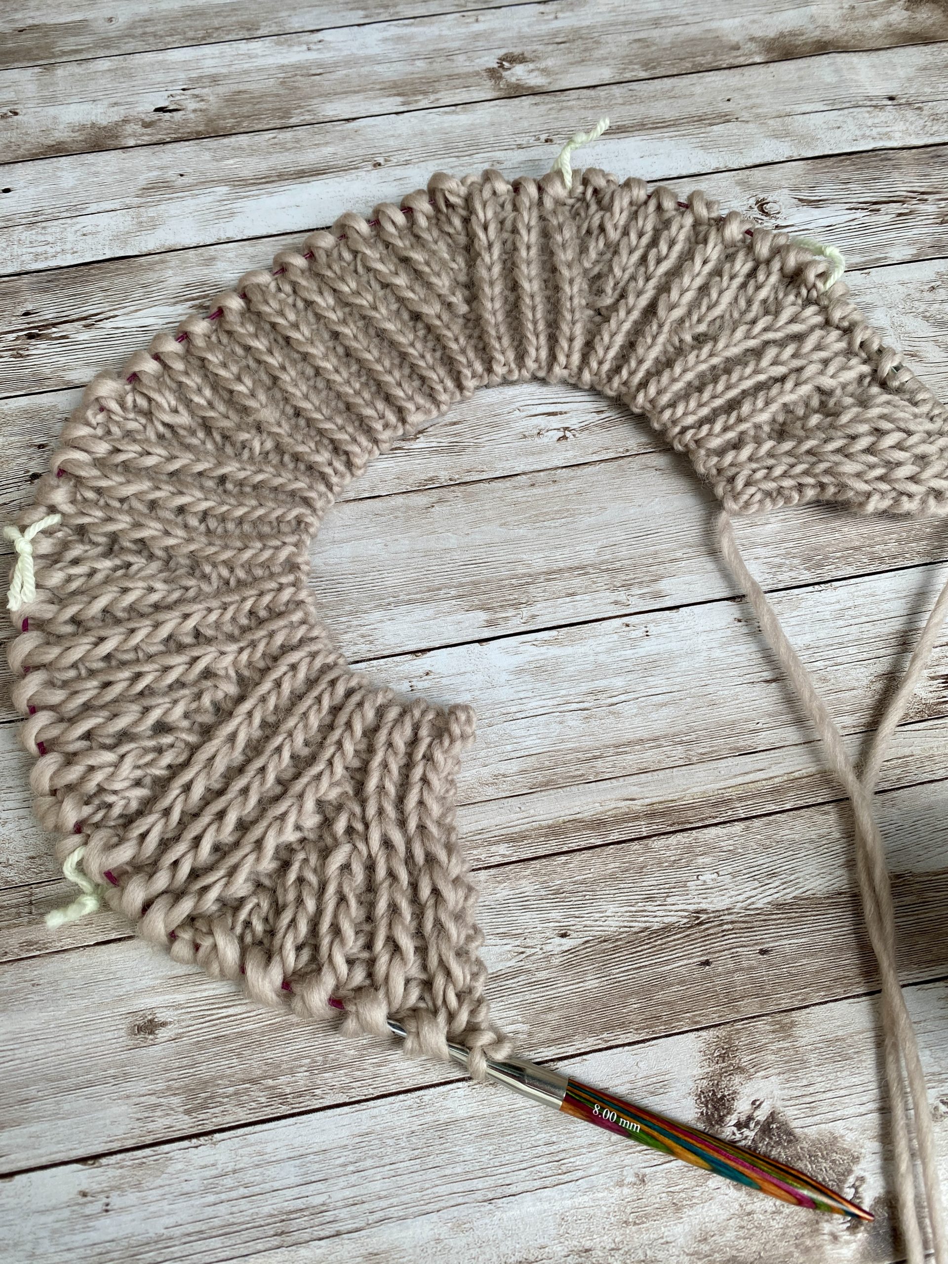 A work-in-progress Fluffy Daisy Cardigan designed by TheKnitStitch, laid flat on a rustic wooden surface. The cardigan is knitted in a chunky beige yarn with a textured ribbed pattern forming a circular shape. Knitting needles are still attached, and a small swatch with embroidered daisies is placed nearby, showcasing a decorative detail for the design.