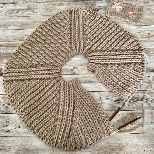 A work-in-progress Fluffy Daisy Cardigan designed by TheKnitStitch, laid flat on a rustic wooden surface. The cardigan is knitted in a chunky beige yarn with a textured ribbed pattern forming a circular shape. Knitting needles are still attached, and a small swatch with embroidered daisies is placed nearby, showcasing a decorative detail for the design.