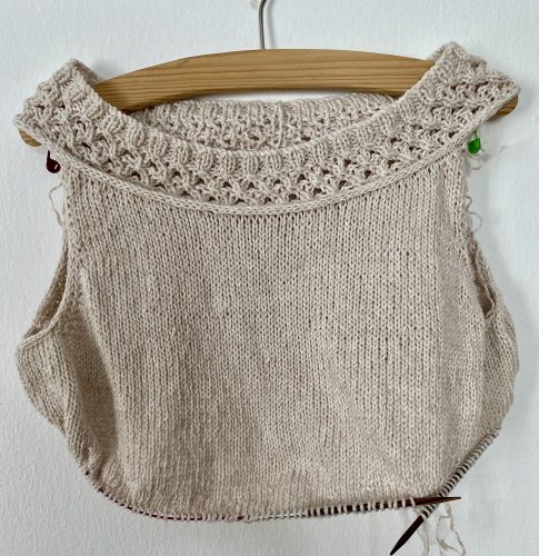 A partially completed Big Chain Top designed by Hinterm Stein, displayed on a wooden hanger against a white wall. The top is knitted in a light beige color, featuring an intricate chain-like lace pattern along the neckline and simple stockinette stitch for the body. The edges are unfinished, with loose yarn strands and knitting needles still attached.