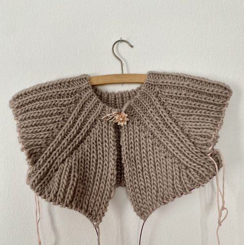 A partially completed Fluffy Daisy Cardigan, designed by TheKnitStitch, displayed on a wooden hanger against a white wall. The cardigan is knitted in a chunky beige yarn with a textured ribbed pattern. A small embroidered daisy detail in soft pink and cream yarn is visible near the neckline. The edges are unfinished, with loose yarn strands and knitting needles still attached.