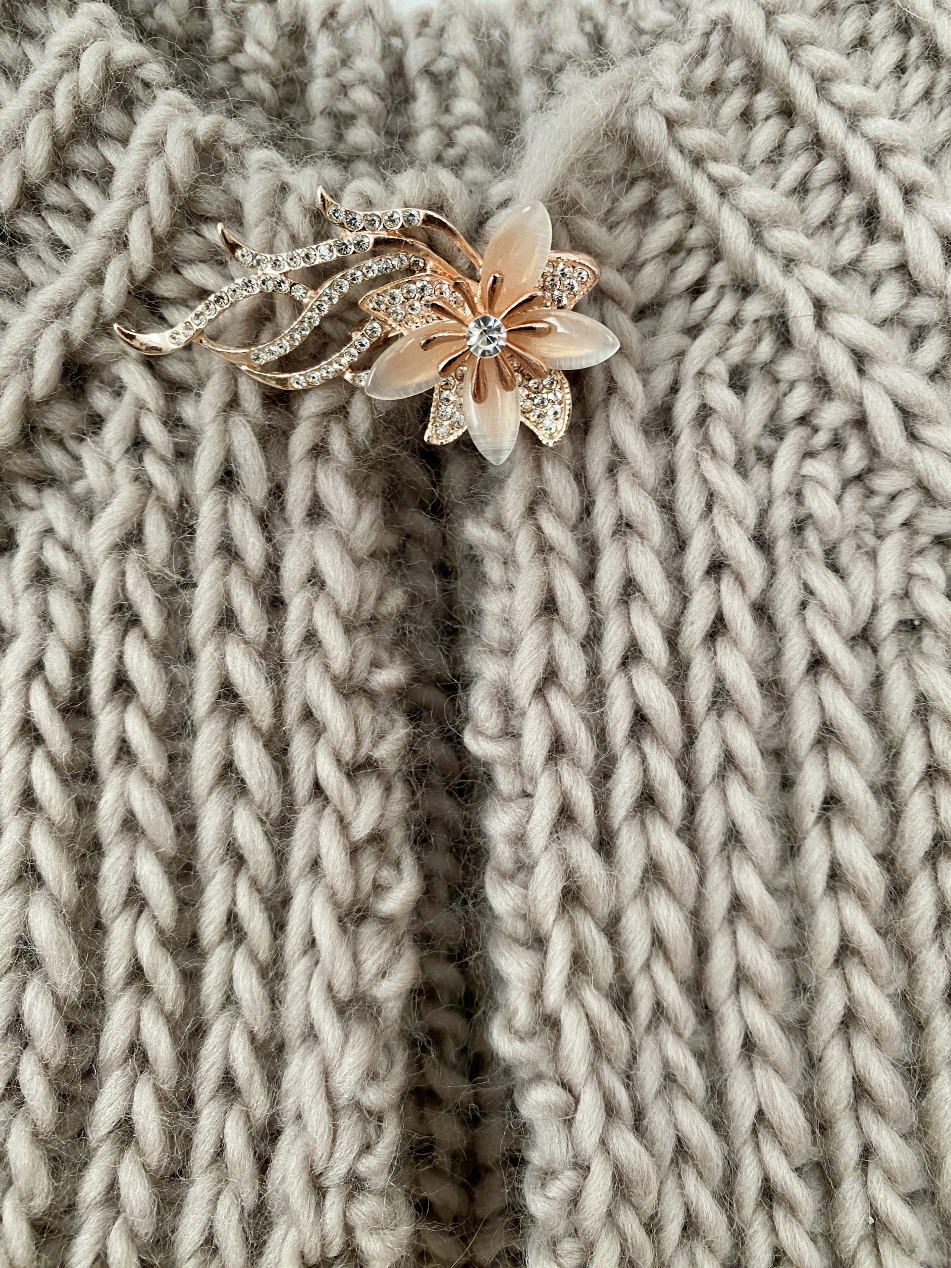 A partially completed Fluffy Daisy Cardigan, designed by TheKnitStitch, displayed on a wooden hanger against a white wall. The cardigan is knitted in a chunky beige yarn with a textured ribbed pattern. A small embroidered daisy detail in soft pink and cream yarn is visible near the neckline. The edges are unfinished, with loose yarn strands and knitting needles still attached.