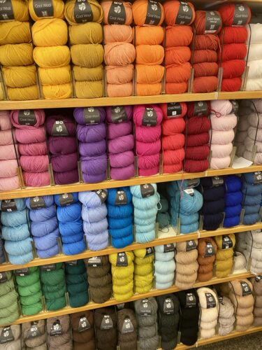 A colorful display of yarn skeins arranged by color on wooden shelves, ranging from warm yellows and reds to cool blues and greens, showcasing a variety of options for knitting projects.