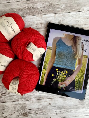 A display of vibrant red yarn skeins labeled Linea Pura Solo Lino next to a tablet showing the Ginkgo Biloba Top pattern. The pattern features a sleeveless, fitted top with a central lace detail, worn by a model holding yellow flowers.