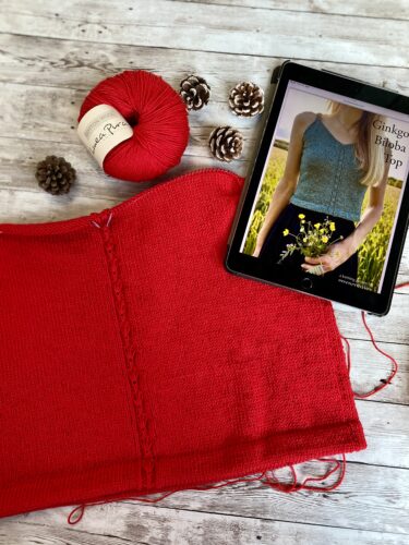 A partially completed Ginkgo Biloba Top in red yarn, laid flat on a rustic wooden surface. The central lace detail is visible, and the tablet displaying the pattern is placed nearby, along with a skein of yarn and decorative pinecones.