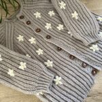The Fluffy Daisy Cardigan, showcasing delicate cream-colored daisy embellishments embroidered onto the chunky beige ribbed knit fabric.