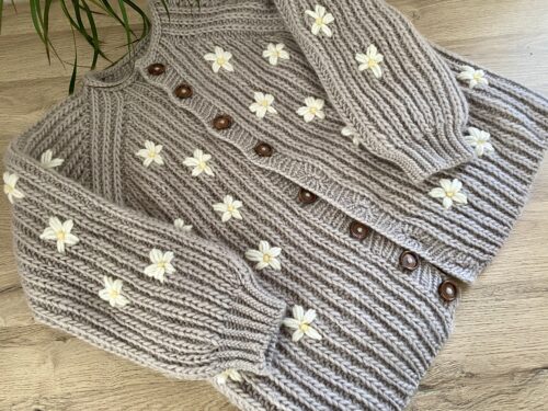 The Fluffy Daisy Cardigan, showcasing delicate cream-colored daisy embellishments embroidered onto the chunky beige ribbed knit fabric.