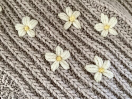 A close-up of the Fluffy Daisy Cardigan, showcasing delicate cream-colored daisy embellishments embroidered onto the chunky beige ribbed knit fabric.