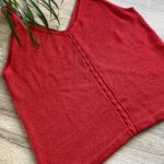 A completed Ginkgo Biloba Top in red yarn, laid flat on a rustic wooden surface. The central lace detail is visible, and the tablet displaying the pattern is placed nearby, along with a skein of yarn and decorative pinecones.