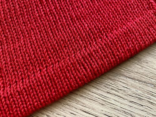 A close-up of the hem of the Ginkgo Biloba Top, showcasing the smooth stockinette stitch in vibrant red yarn, laid on a wooden surface.