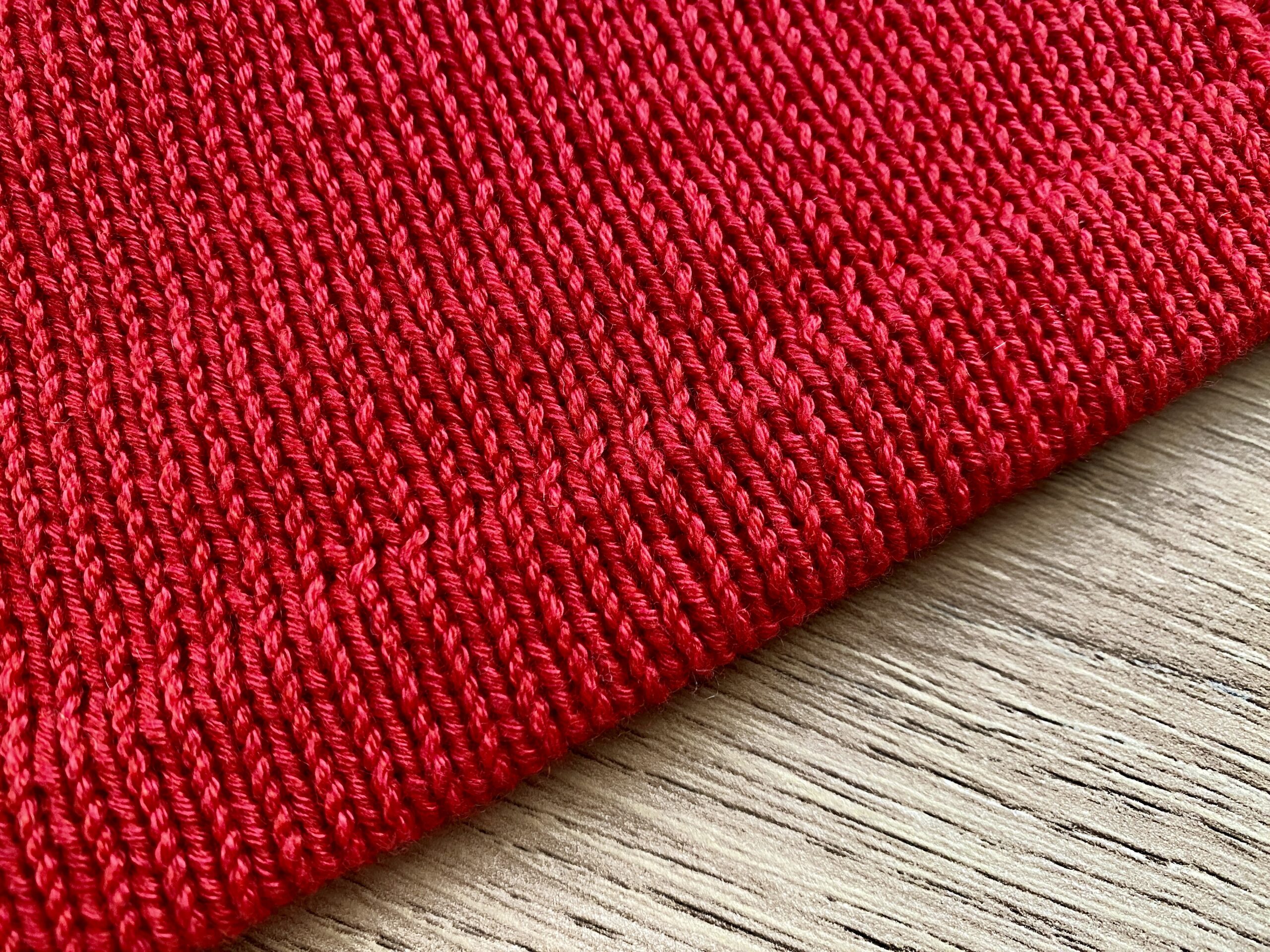 A close-up of the hem of the Ginkgo Biloba Top, showcasing the smooth stockinette stitch in vibrant red yarn, laid on a wooden surface.