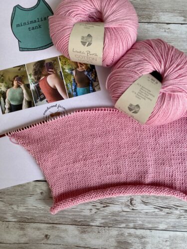 A partially knitted Minimalist Tank Top in soft pink yarn, displayed with two skeins of 