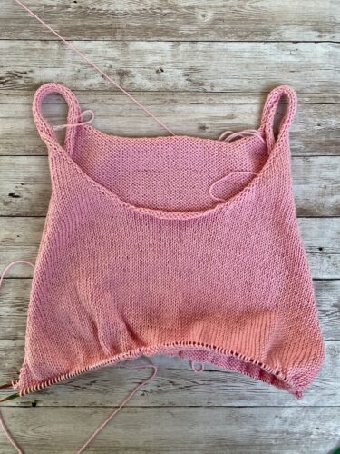 A work-in-progress Minimalist Tank Top in soft pink yarn, laid flat on a rustic wooden surface. The tank top features a simple stockinette stitch with thin straps and a scooped neckline.