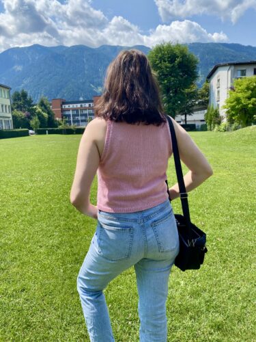 A back view of the Minimalist Tank Top in soft pink, worn by a woman standing in a grassy field with mountains and buildings in the background. The tank top is paired with light blue jeans and a black crossbody bag.