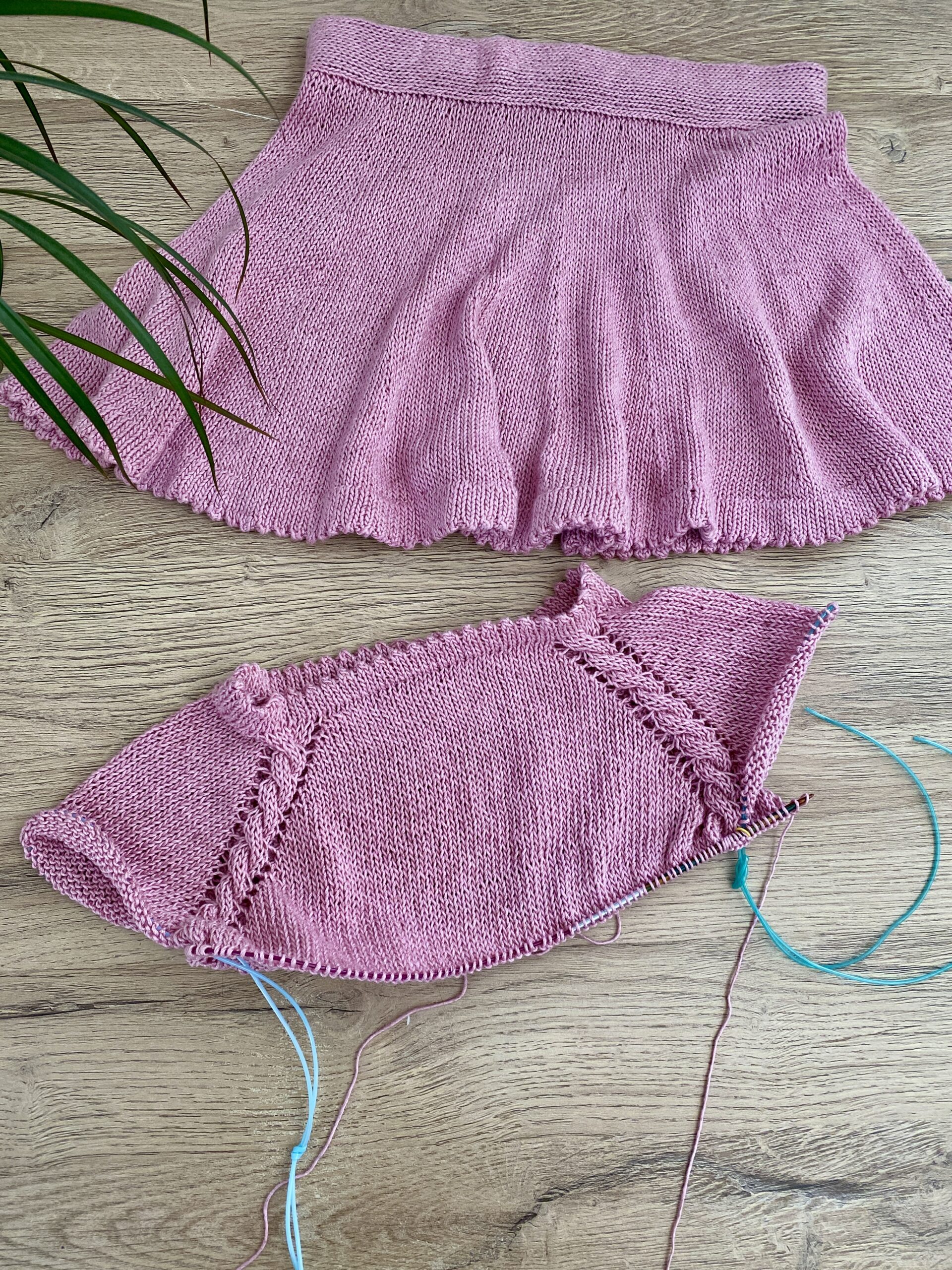 The Skeeter Skirt and a matching top in progress, both in soft pink yarn. The flared skirt is completed, while the top is partially knitted, with loose yarn strands and needles visible.