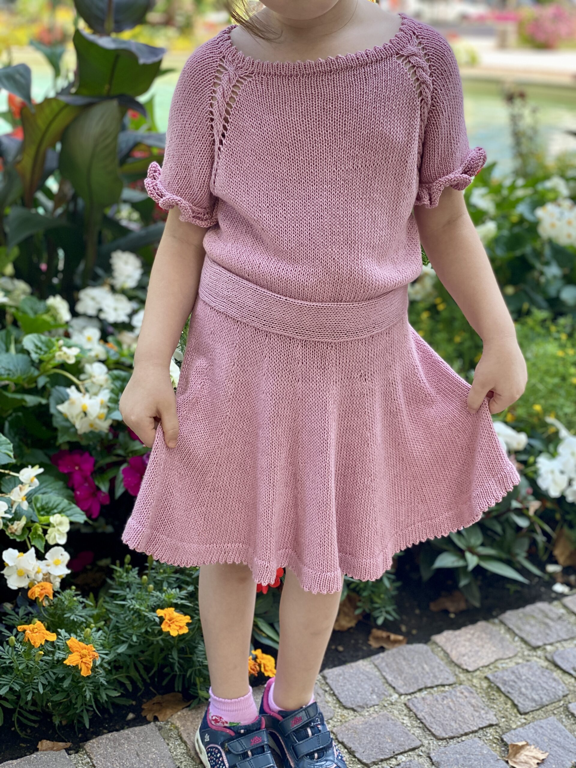 A child wearing the Skeeter Skirt and matching top in soft pink, standing in a garden surrounded by colorful flowers. The skirt features a flared design with a delicate hem, paired with a short-sleeved top with ruffled edges.
