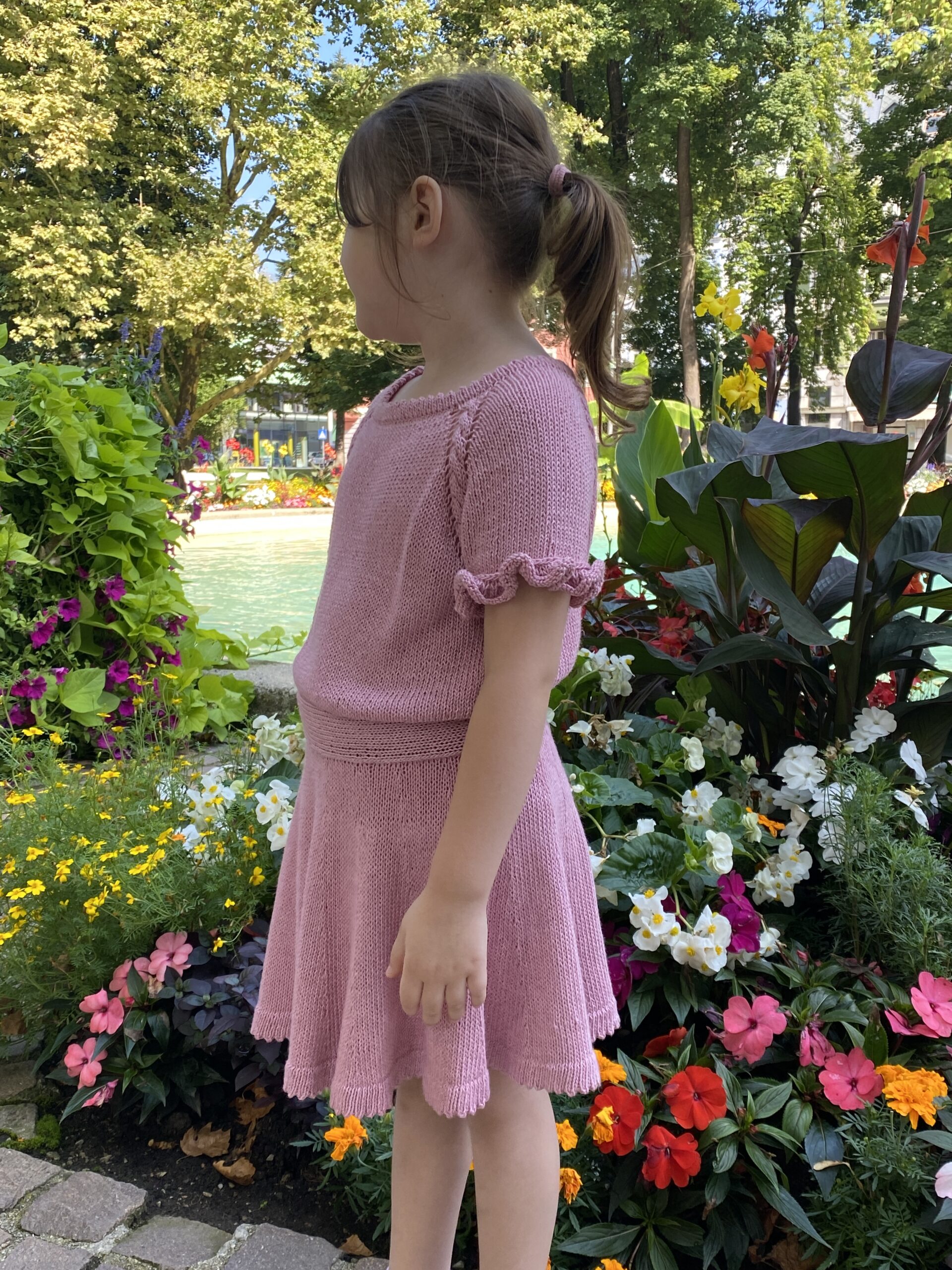 A side view of a child wearing the Skeeter Skirt and matching top in soft pink, standing in a vibrant garden. The flared skirt and ruffled short sleeves add a playful and elegant touch to the outfit.