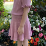 A child wearing the Skeeter Skirt and matching top in soft pink, standing in a garden surrounded by colorful flowers. The skirt features a flared design with a delicate hem, paired with a short-sleeved top with ruffled edges.