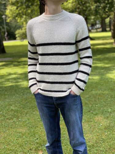A front view of the Vine Pullover, showcasing its classic cream and black striped design with a crew neckline and a relaxed fit. The pullover is worn with blue jeans in a sunny park setting.