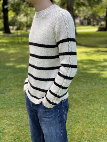 A side view of the Vine Pullover, worn by a person standing in a grassy park. The pullover is cream-colored with black horizontal stripes and features a subtle cable detail on the sleeves, paired with blue jeans.