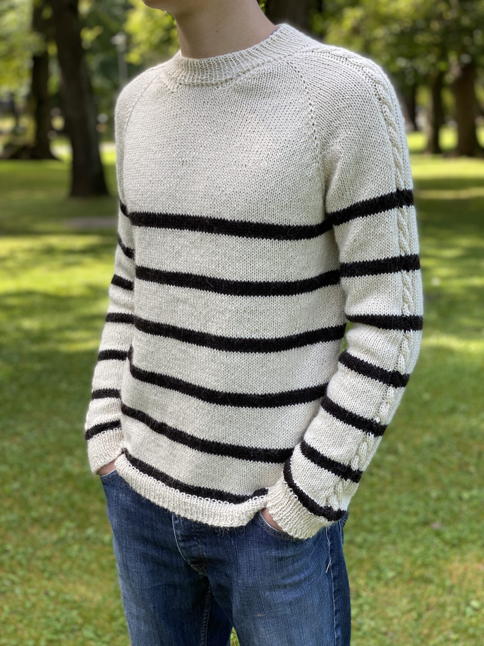 A side view of the Vine Pullover, worn by a person standing in a grassy park. The pullover is cream-colored with black horizontal stripes and features a subtle cable detail on the sleeves, paired with blue jeans.