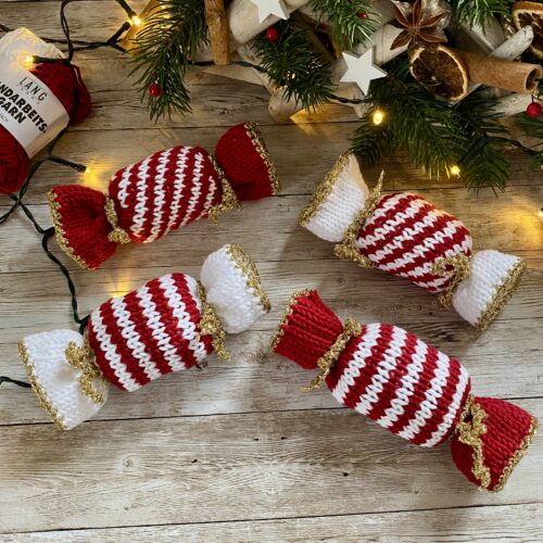 A festive display of knitted Christmas Caramels in red and white stripes with gold accents, arranged on a rustic wooden surface. The caramels are surrounded by holiday decorations, including greenery, cinnamon sticks, and warm string lights.