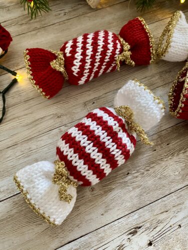 A mix of red and white striped knitted Christmas Caramels with gold accents, arranged on a wooden surface. The festive decorations include greenery and warm holiday lights.