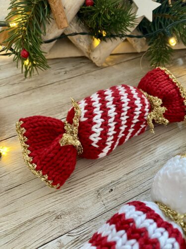 A close-up of a red and white striped knitted Christmas Caramel with gold trim and bow details, placed on a wooden surface. Holiday greenery and lights are visible in the background.