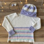 A hand-knitted cream sweater with pastel-colored stripes and lavender ribbed cuffs, paired with a matching lavender and cream striped hat. Knitting tools and yarn are displayed in the background on a wooden surface with fairy lights.