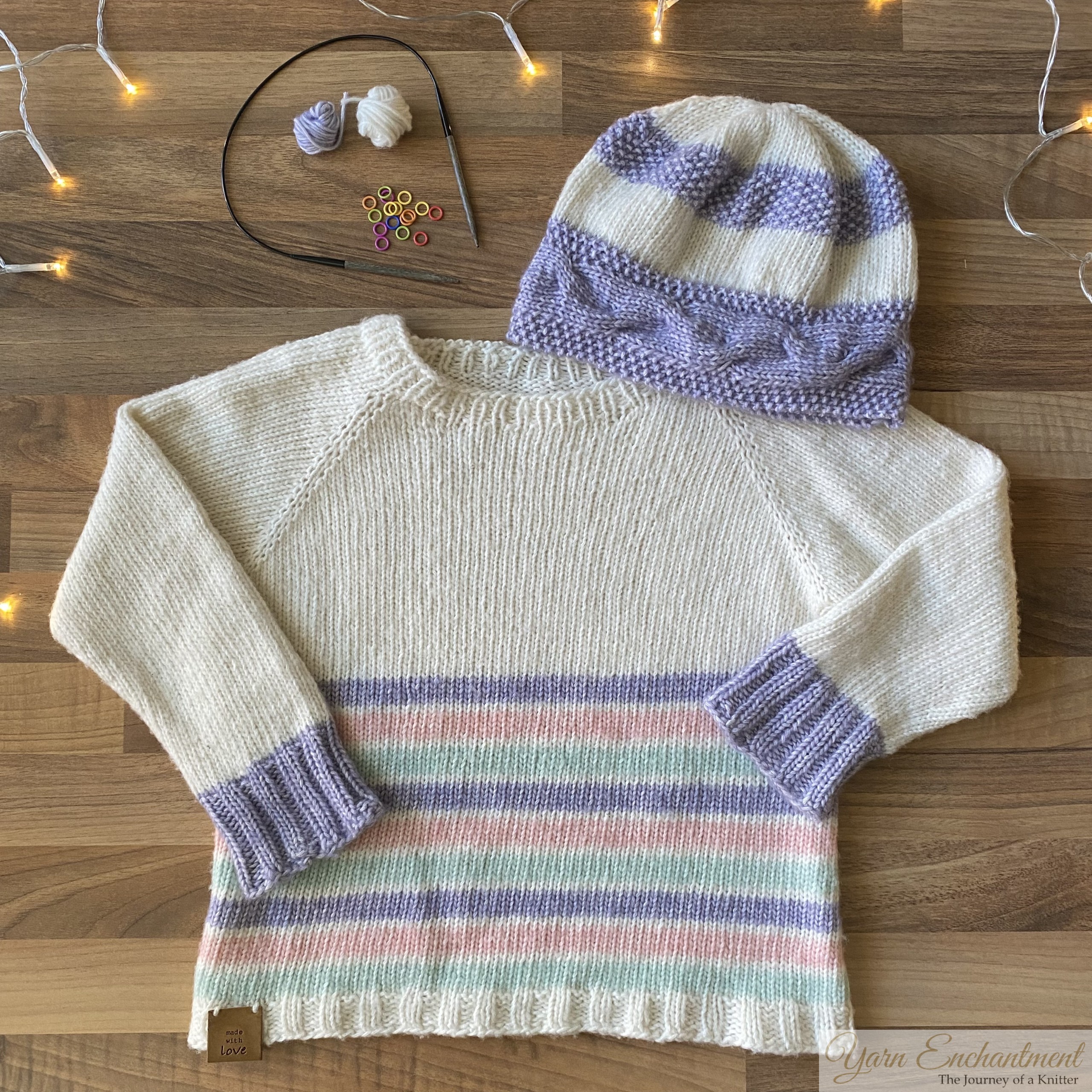 A hand-knitted cream sweater with pastel-colored stripes and lavender ribbed cuffs, paired with a matching lavender and cream striped hat. Knitting tools and yarn are displayed in the background on a wooden surface with fairy lights.
