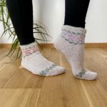 A person wearing hand-knitted, light beige ankle socks with floral patterns in pink, green, and purple. The socks have a scalloped edge at the cuff and a decorative green vine design near the toes. The person is standing on a wooden floor, with a green plant in the background.