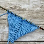 Close-up of knitted swatch on wooden knitting needles, in blue yarn, showing the make one left and right increases