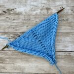 Close-up of knitted swatch on wooden knitting needles, in blue yarn, showing the make one left and right increases on the purl side