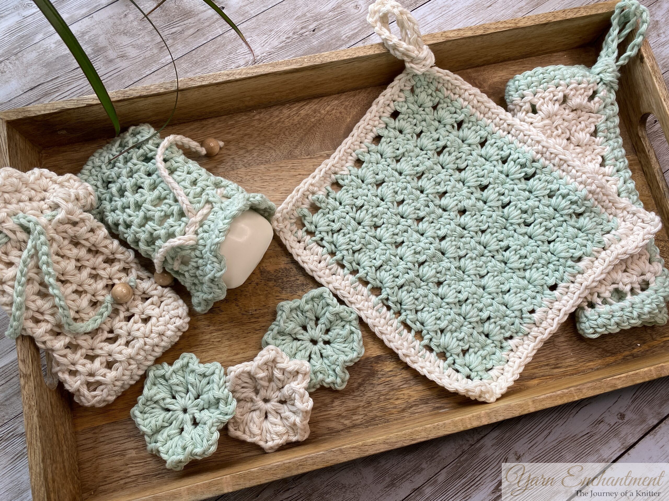 A handmade crochet washcloth in soft mint green with a white border, featuring the Primrose Stitch. The washcloth has a built-in hanger loop tied at the top and is displayed on a wooden tray alongside matching crochet pieces.