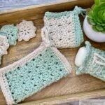 A collection of handmade crochet items in mint green and white, including washcloths, floral motifs, and a soap pouch with a drawstring. The items are arranged on a wooden tray with a small succulent plant for decoration.