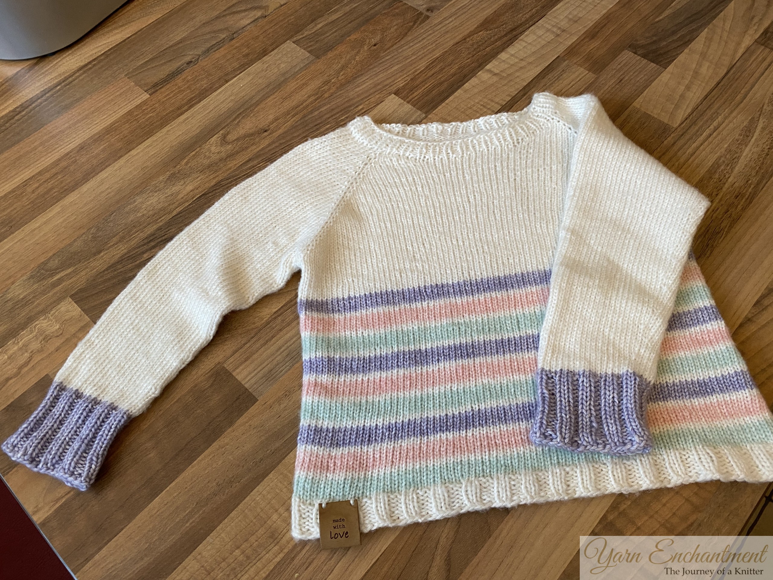 A hand-knitted cream sweater with pastel-colored stripes and lavender ribbed cuffs, laid flat on a wooden surface, showcasing its simple yet elegant design.