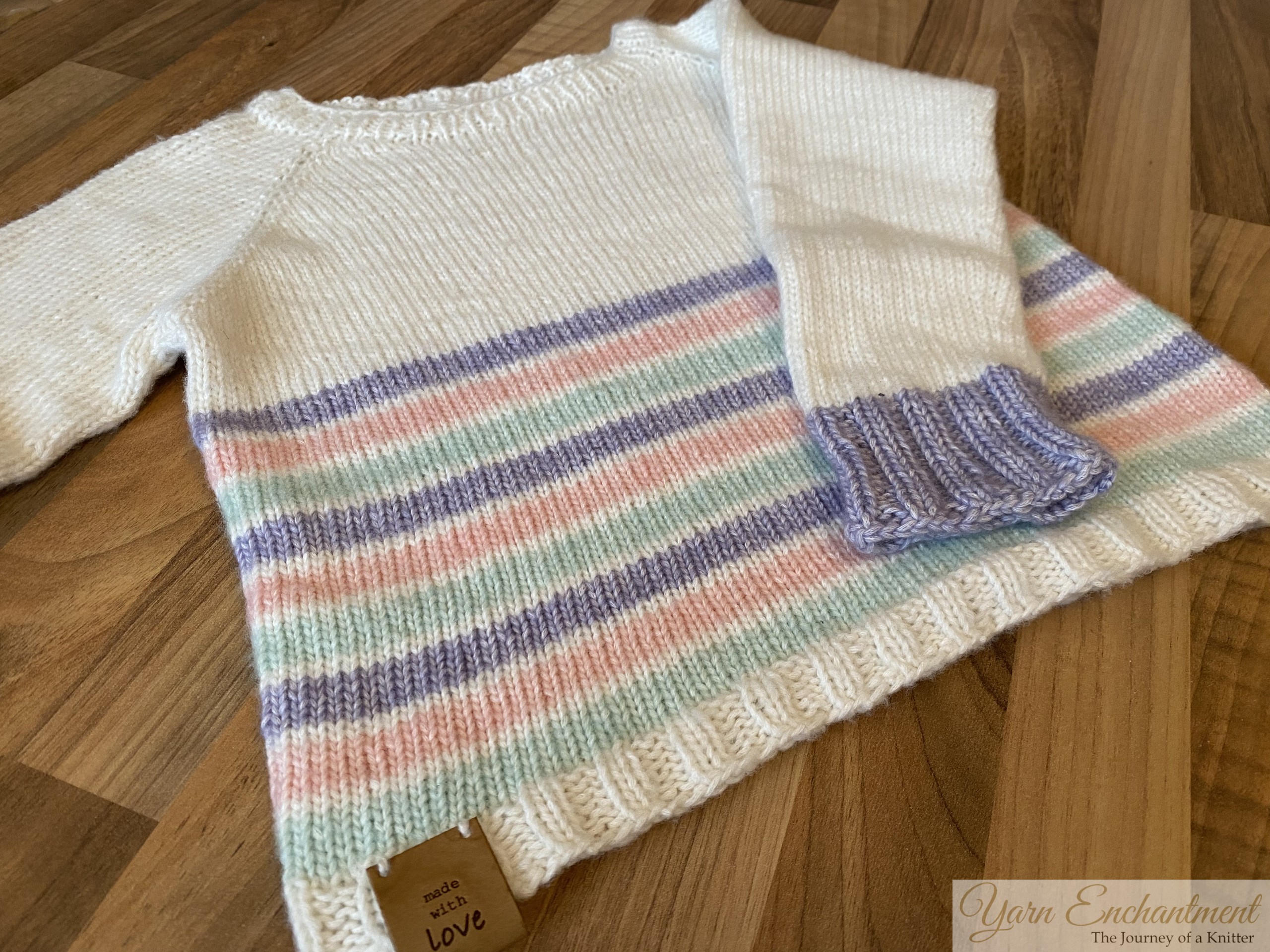 A hand-knitted cream sweater with pastel-colored stripes and lavender ribbed cuffs, laid flat on a wooden surface, showcasing its simple yet elegant design.