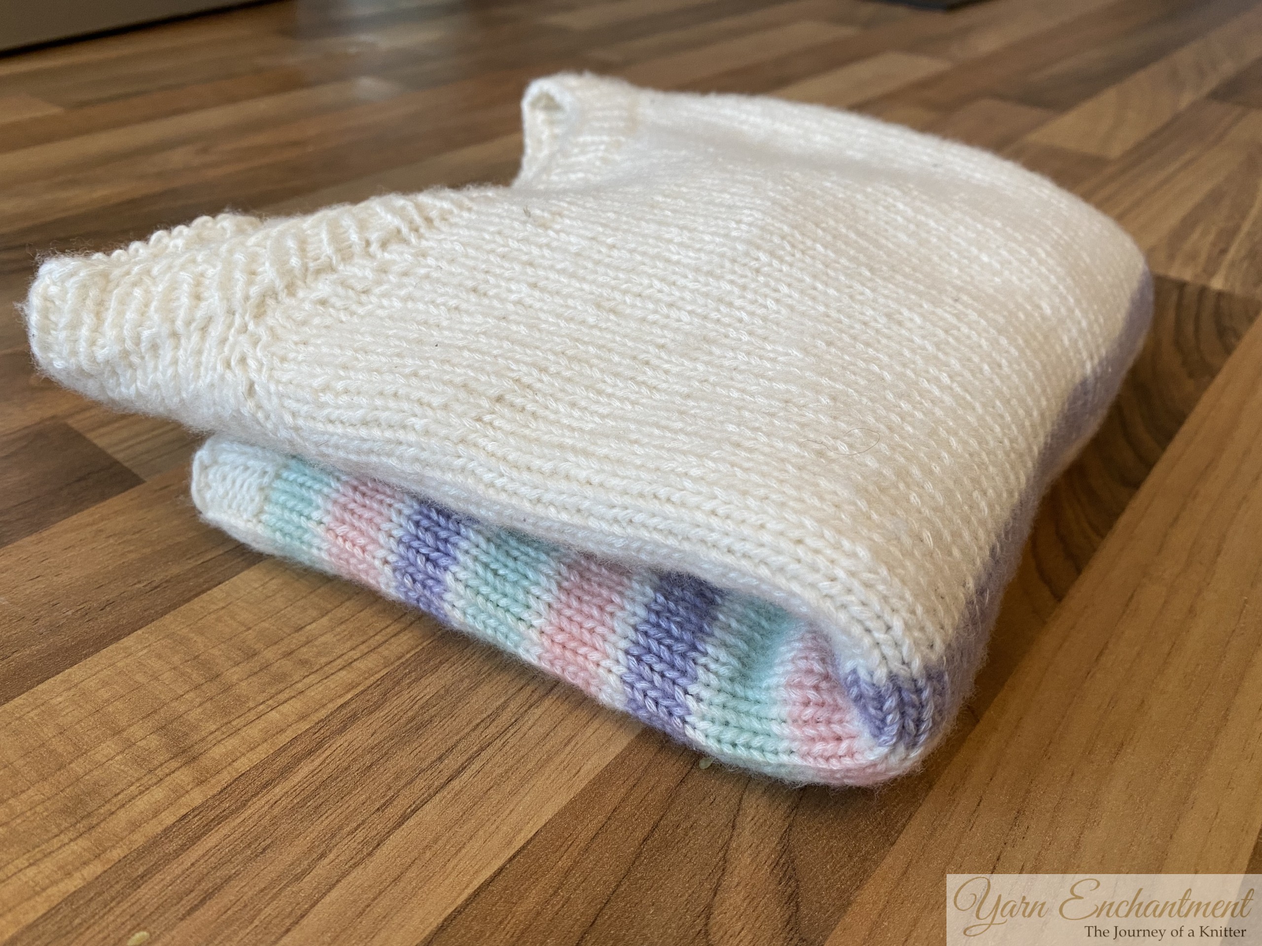 A hand-knitted cream sweater with pastel-colored stripes and lavender ribbed cuffs, laid flat on a wooden surface, showcasing its simple yet elegant design.
