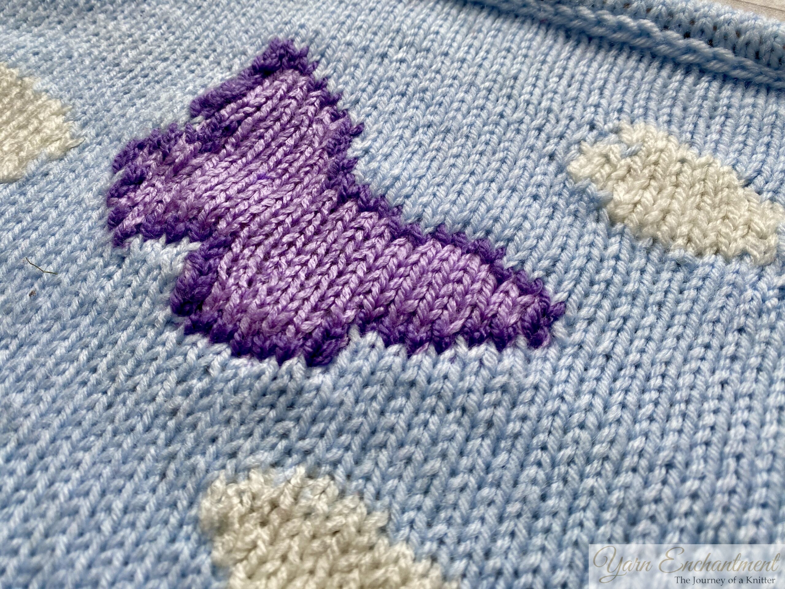 A knitted square using the intarsia technique, depicting a landscape with green hills, two trees with red apples, white clouds, and a purple butterfly against a light blue sky.
