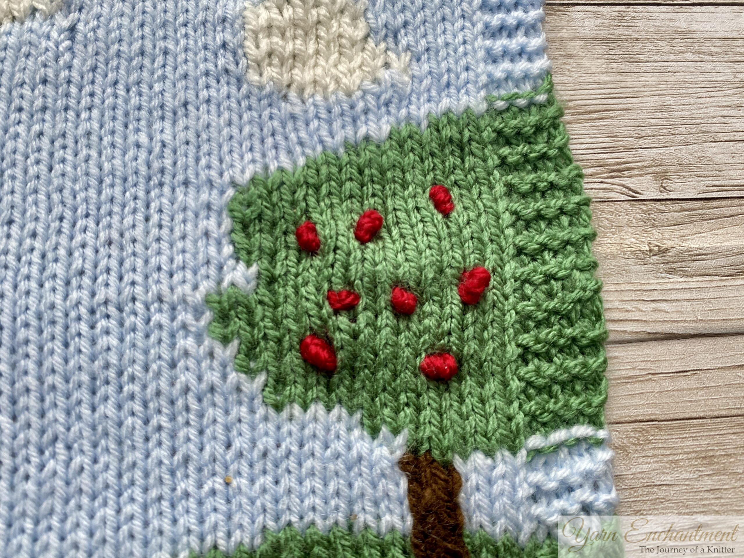 A knitted square using the intarsia technique, depicting a landscape with green hills, two trees with red apples, white clouds, and a purple butterfly against a light blue sky. The little apples are french knots