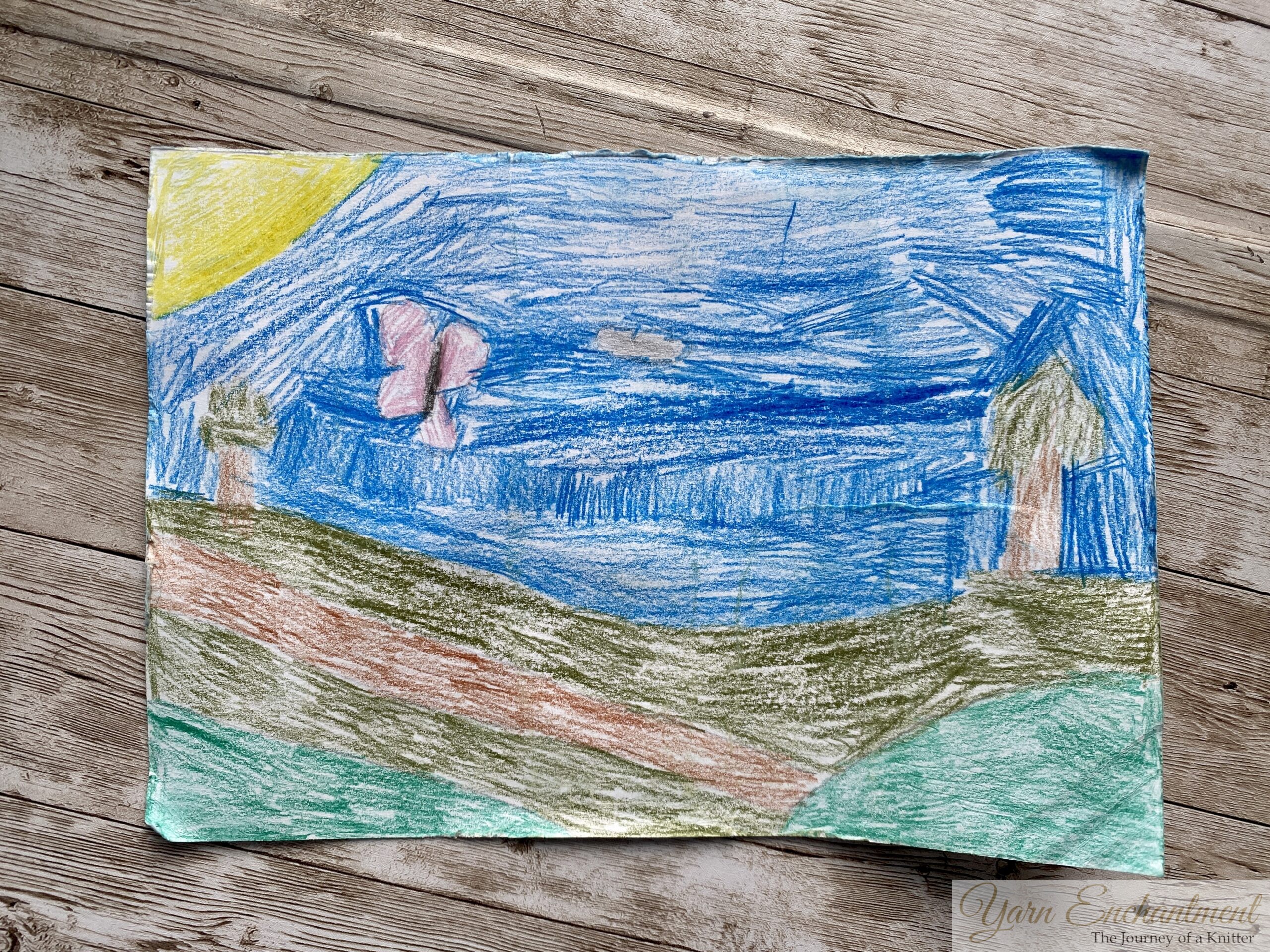 A child's crayon drawing of a landscape with a blue sky, green hills, two trees, and a pink butterfly, with a yellow sun in the corner.