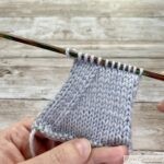 A small triangular knitted swatch on a wooden needle, showing the result of "k2tog" decreases along the edges, creating a symmetrical shape.