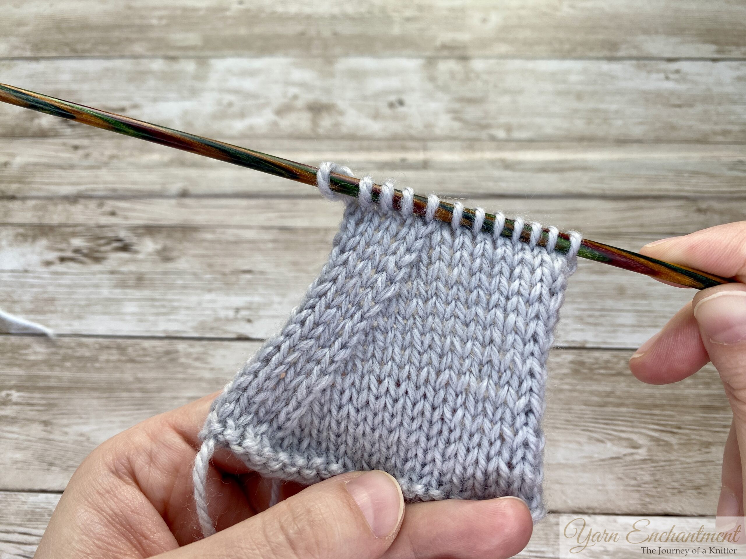A small triangular knitted swatch on a wooden needle, showing the result of 'k2tog' decreases along the edges, creating a symmetrical shape.