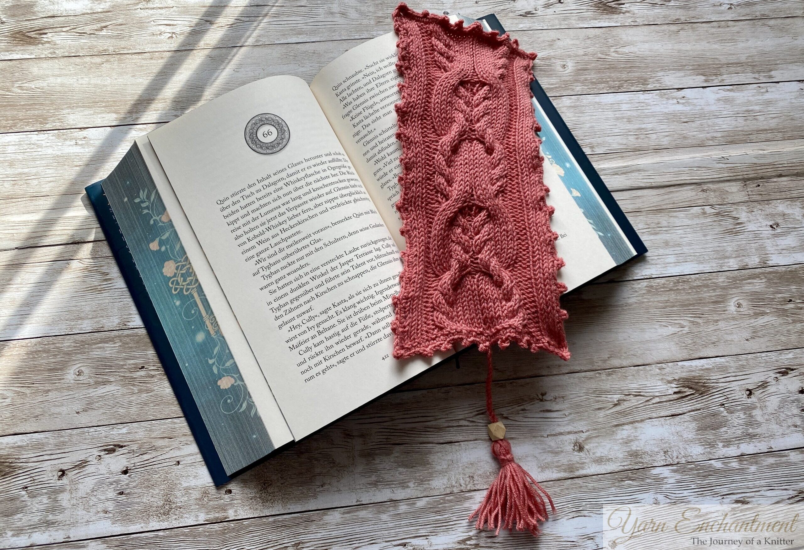 An open book with a knitted pink bookmark placed across the pages. The bookmark features intricate cable patterns and a scalloped edge, lying flat on the book, which is set on a rustic wooden surface.