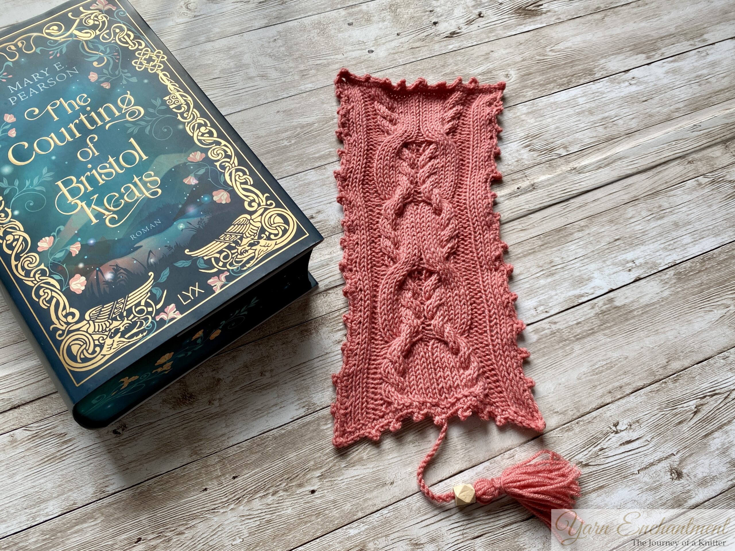 A beautifully knitted pink bookmark with intricate cable patterns lies next to the book 'The Courting of Bristol Keats' by Mary E. Pearson. The book has an elegant dark teal cover with gold floral and decorative accents. The bookmark features a tassel at the bottom and is placed on a rustic wooden surface, creating a cozy and creative atmosphere.