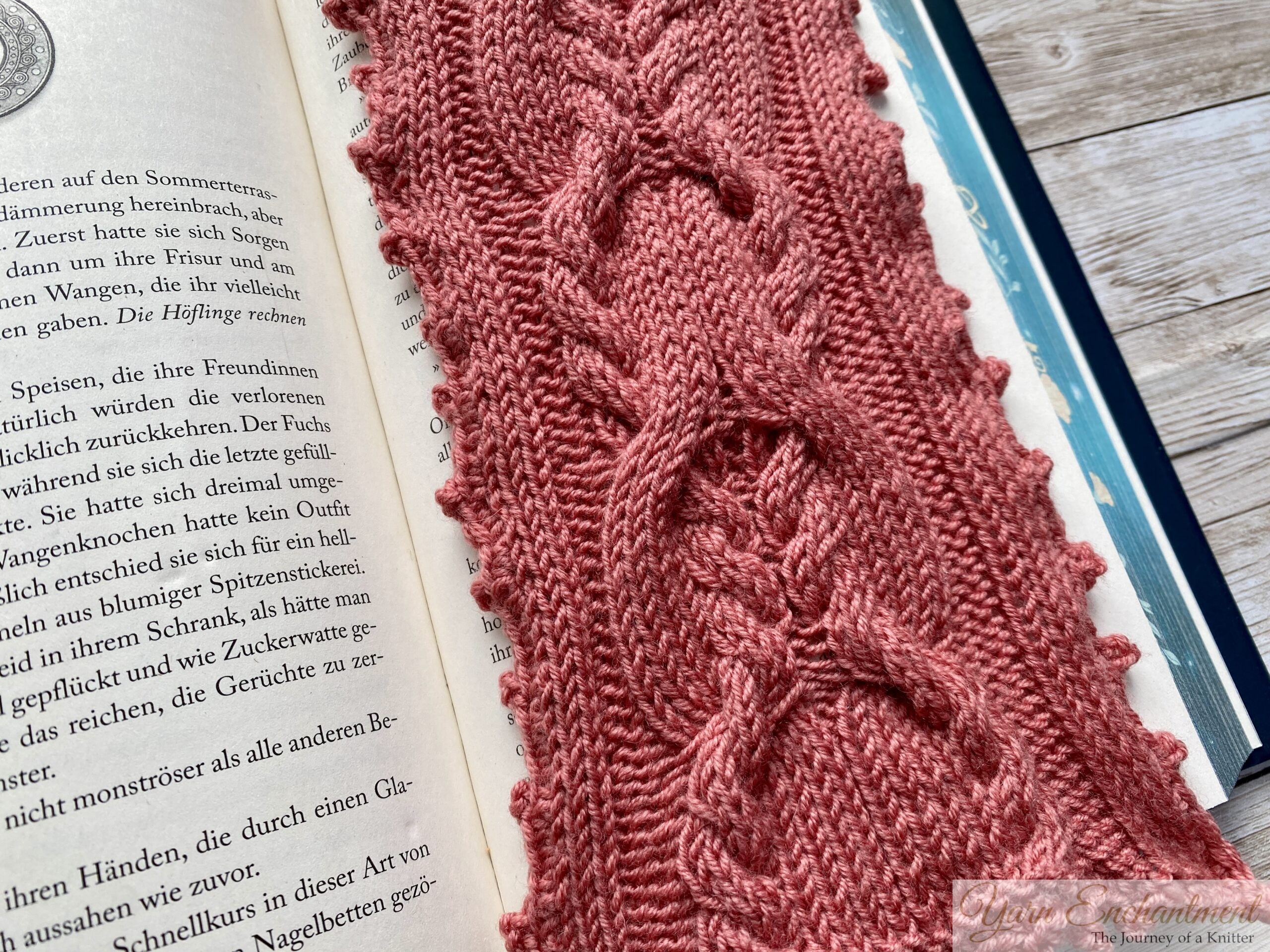 A detailed close-up of a pink knitted bookmark with elegant cable patterns and a scalloped edge. The bookmark is placed on an open book, showcasing its intricate design against the printed text.