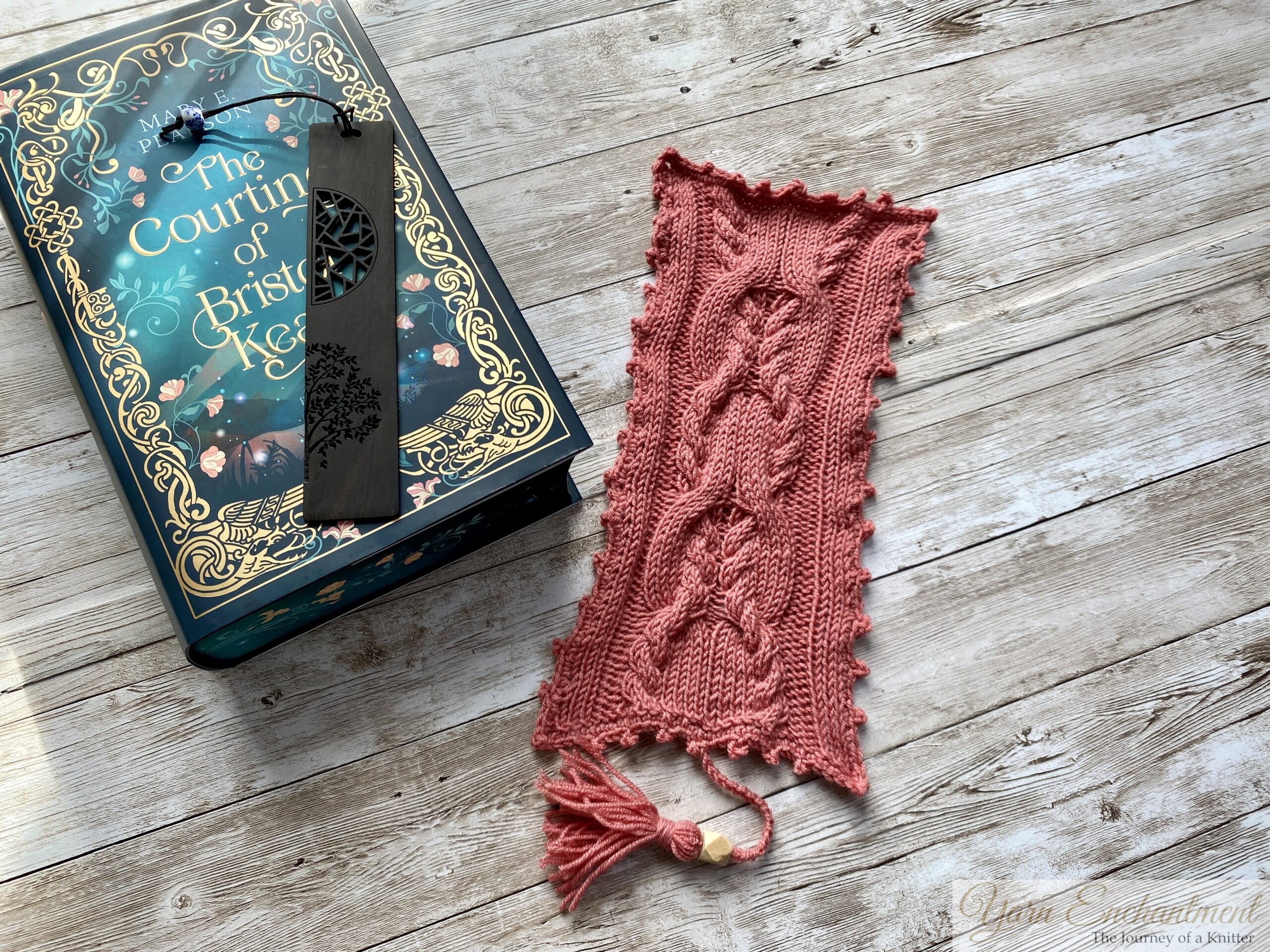 A pink knitted bookmark with intricate cable patterns and a scalloped edge lies next to the book 'The Courting of Bristol Keats' by Mary E. Pearson. The book has a dark teal cover with gold floral accents, and a black metal bookmark with a tree design is placed on top of it. Both items rest on a rustic wooden surface, creating a cozy and creative scene.