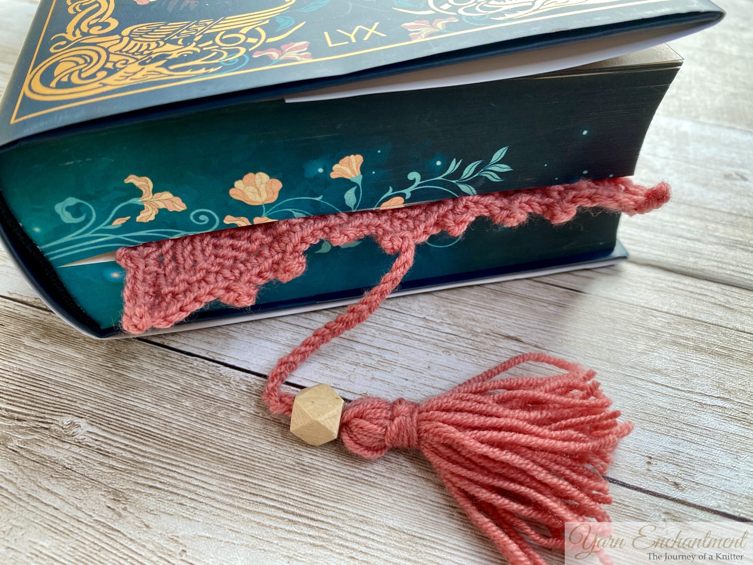 A close-up of a knitted pink bookmark with a scalloped edge and a tassel, placed inside a dark teal book with gold floral accents on the cover. The bookmark hangs out of the book, resting on a rustic wooden surface