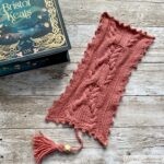 A beautifully knitted pink bookmark with intricate cable patterns lies next to the book 'The Courting of Bristol Keats' by Mary E. Pearson. The book has an elegant dark teal cover with gold floral and decorative accents. The bookmark features a tassel at the bottom and is placed on a rustic wooden surface, creating a cozy and creative atmosphere.