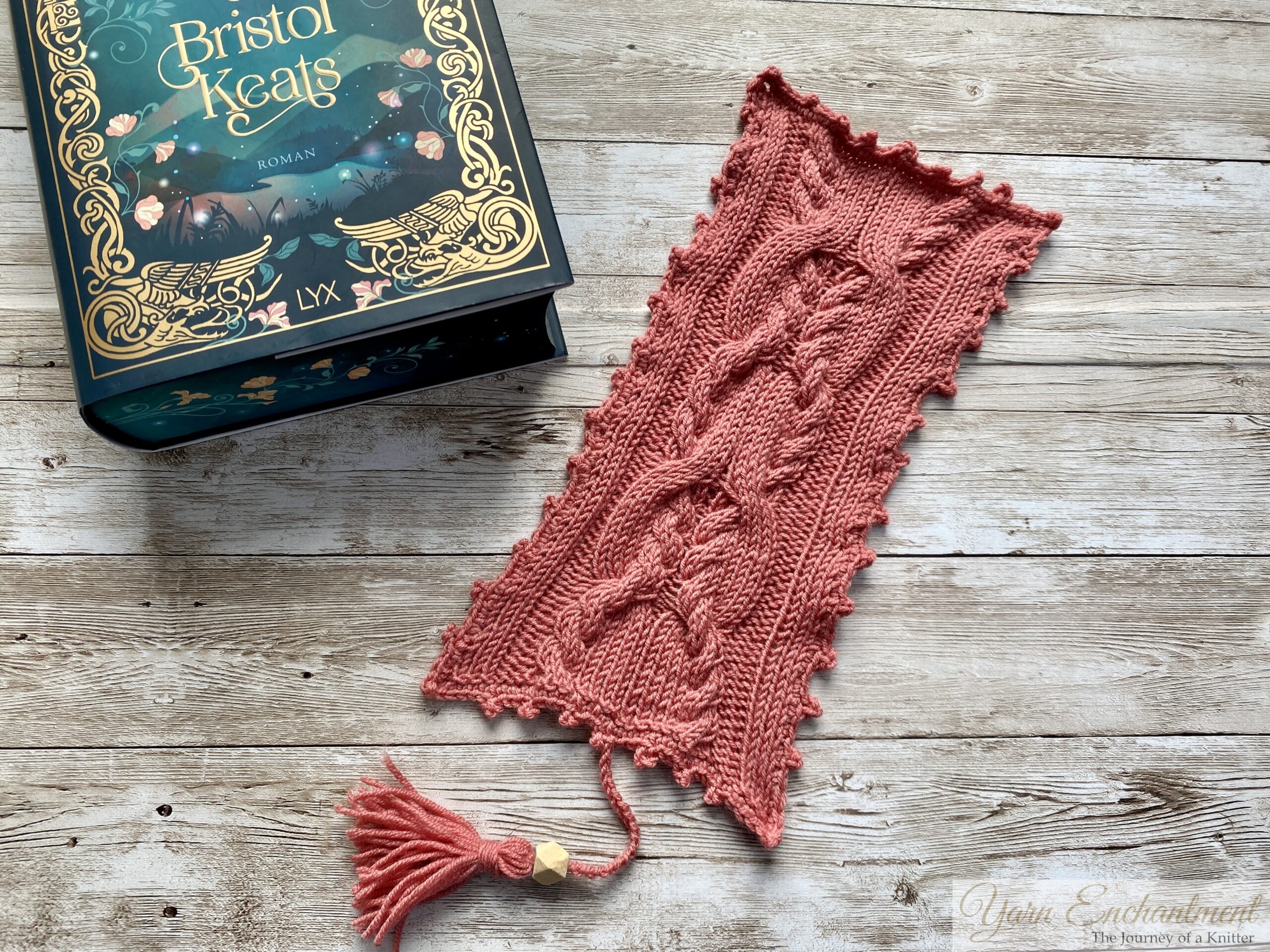 A beautifully knitted pink bookmark with intricate cable patterns lies next to the book 'The Courting of Bristol Keats' by Mary E. Pearson. The book has an elegant dark teal cover with gold floral and decorative accents. The bookmark features a tassel at the bottom and is placed on a rustic wooden surface, creating a cozy and creative atmosphere.