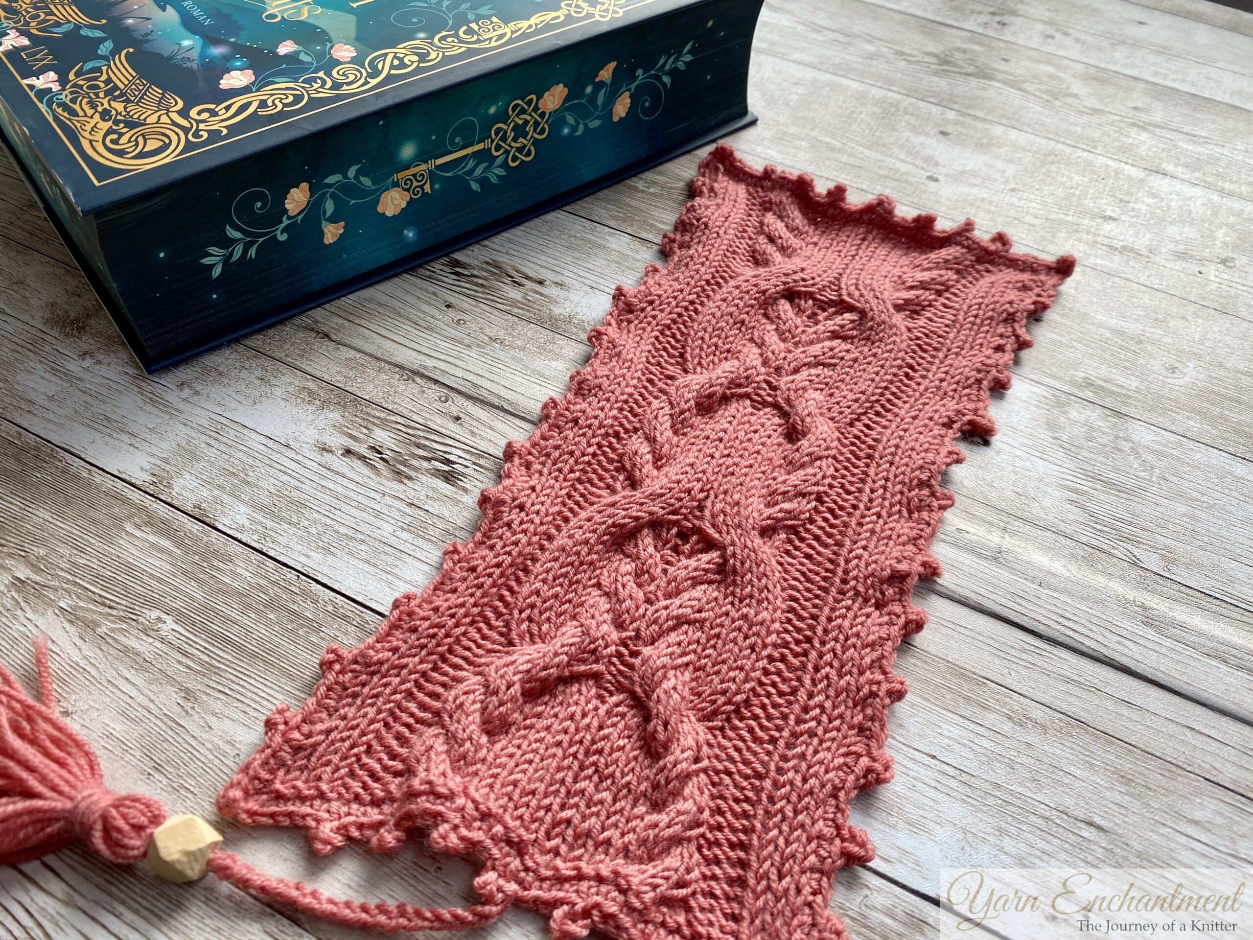A close-up view of a pink knitted bookmark showcasing its intricate cable patterns and scalloped edges. The bookmark is placed on a wooden surface, with part of a book visible in the corner.